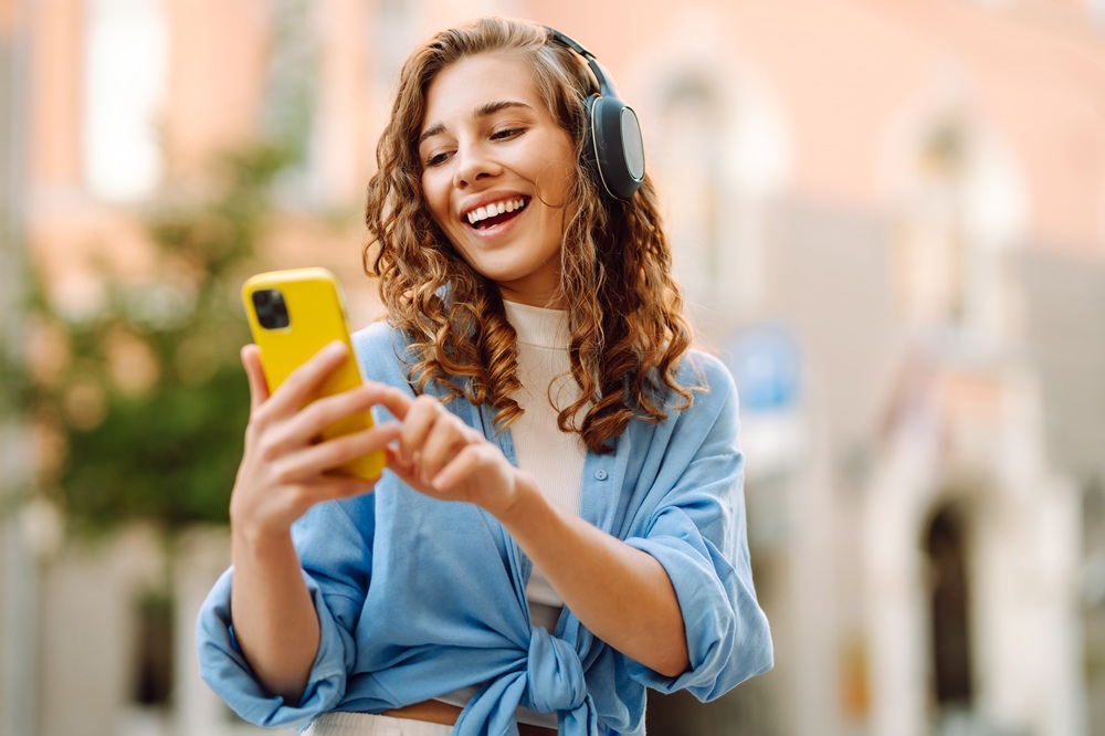 Smiling young woman listening to music on headphones and using smartphone – Dentist Wenatchee WA Cheerful woman wearing headphones and holding a yellow smartphone while enjoying music outdoors – Dentist Wenatchee WA
