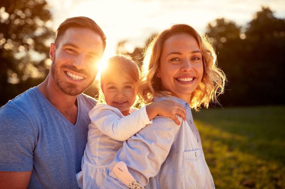 Happy family smiling together at sunset – Dentist Wenatchee WA Smiling parents holding their young daughter while enjoying a warm sunset in a green park, symbolizing healthy family smiles – Dentist Wenatchee WA